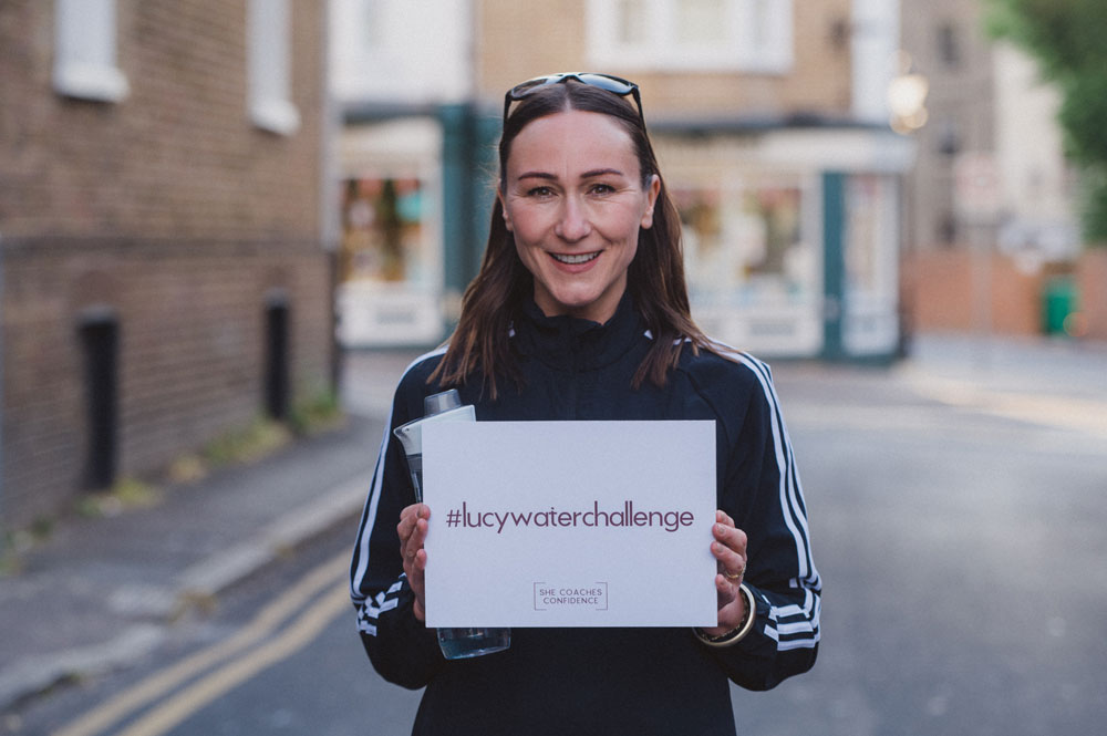 Lucy Baker standing holding a sign saying #lucywaterchallenge and wearing a black adidas top
